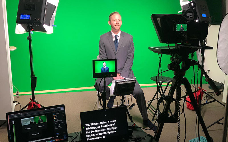 Behind the scenes shot showing businessman reading off a teleprompter while sitting in front of a greenscreen